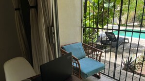 Una habitación con una computadora portátil sobre una mesa de madera, una silla de mimbre con un cojín azul y vistas a una piscina a través de una reja metálica.