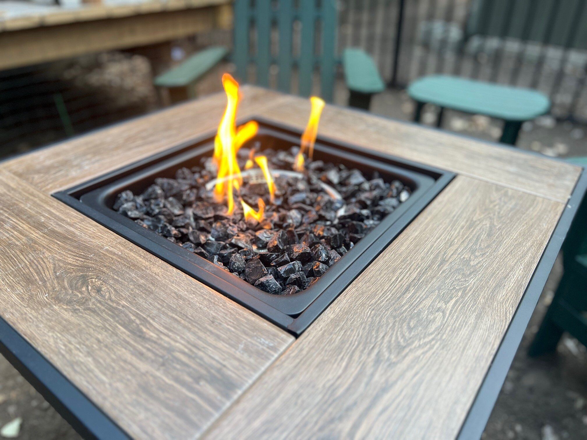 A square fire pit with burning logs, surrounded by a wooden table and chairs.