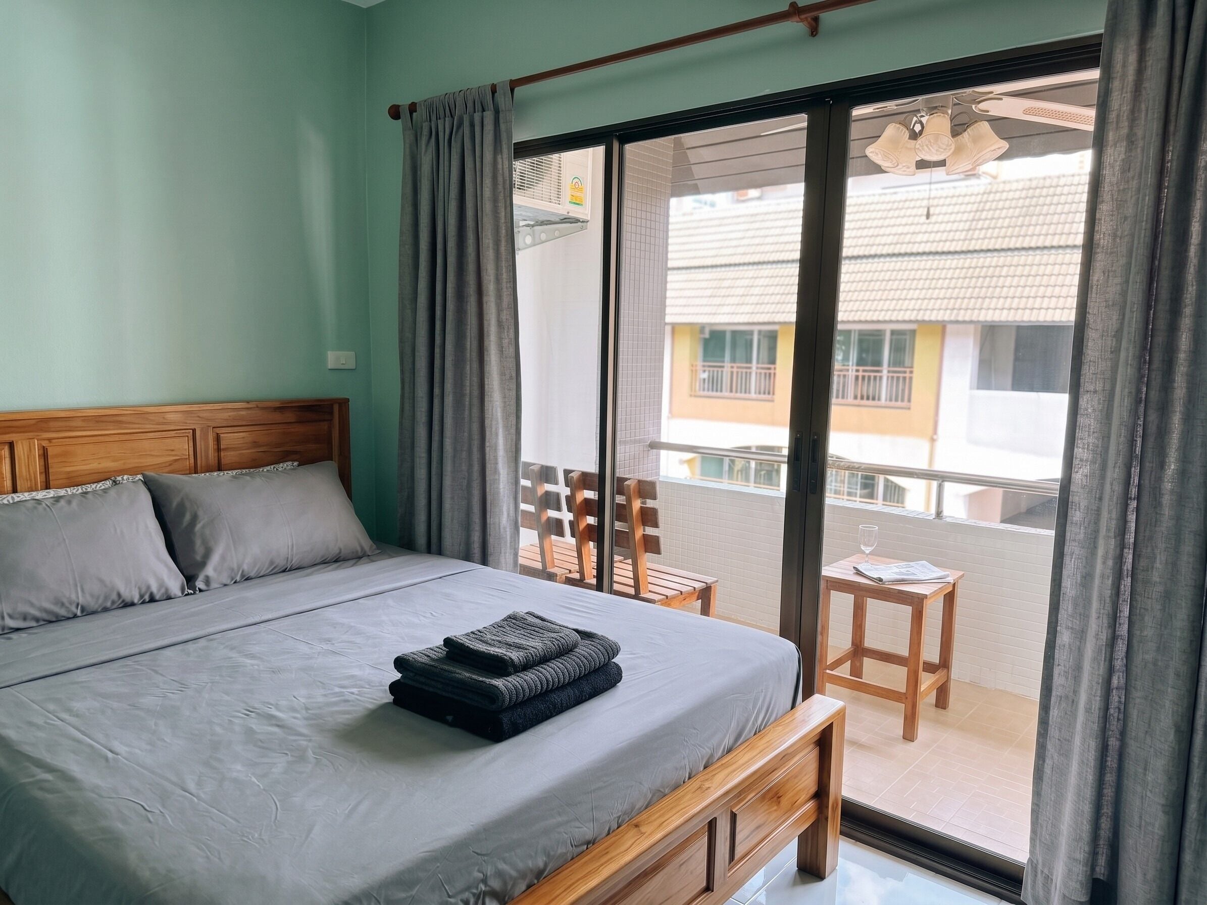 Une chambre à coucher avec un lit, une tête de lit en bois, un balcon avec une table et des chaises, et une vue sur les bâtiments environnants.