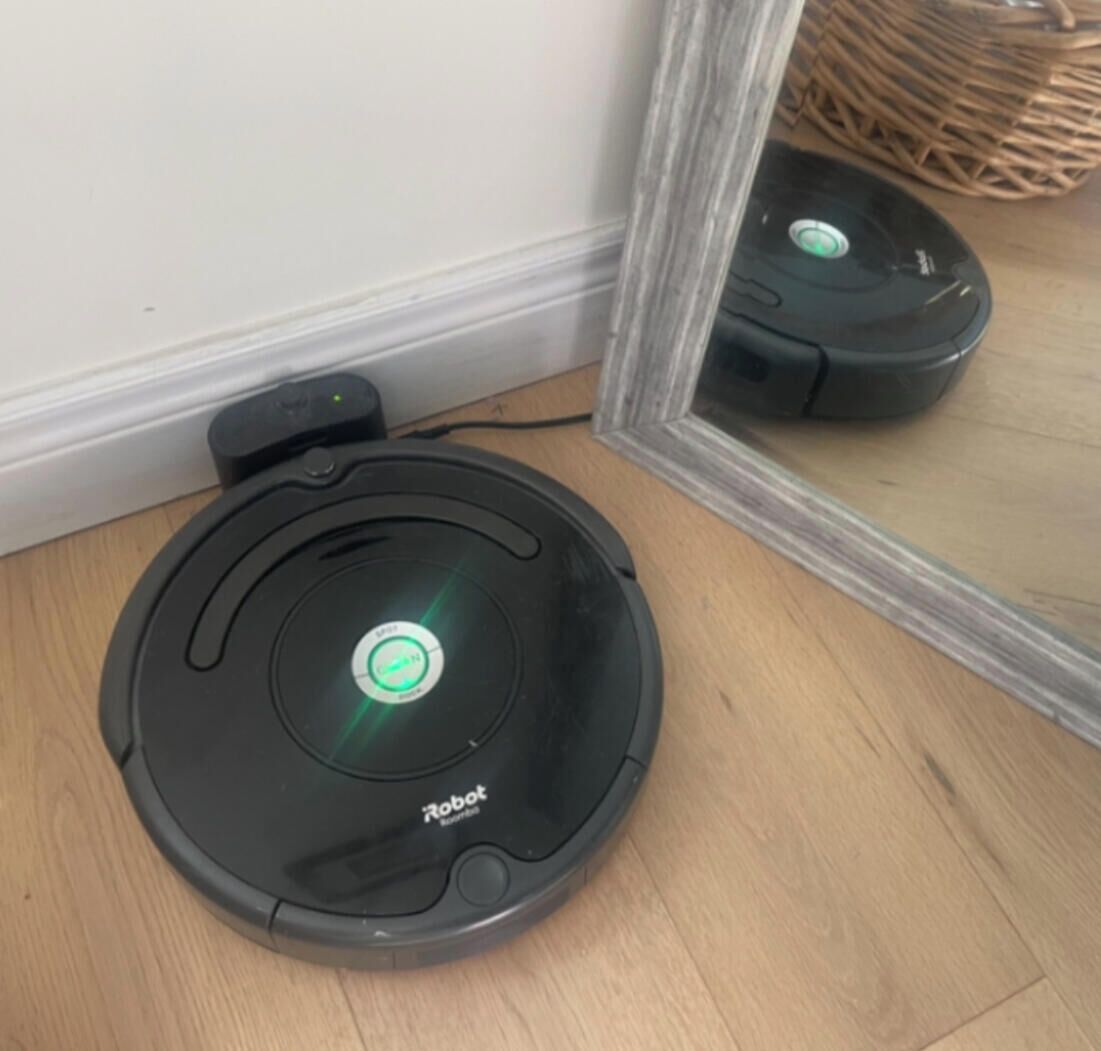 Un robot aspirador en una habitación con suelo de madera y un espejo que refleja parte del espacio.