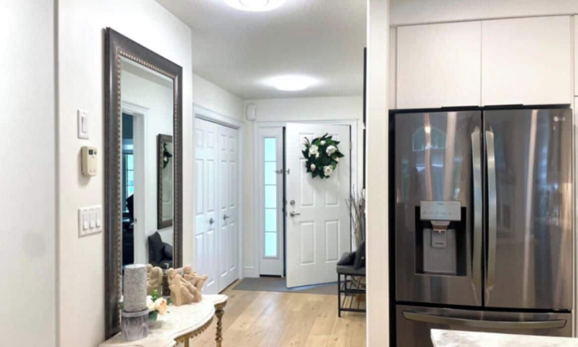 A hallway with a mirror, a door with a wreath, a table with a vase, and a stainless steel refrigerator.