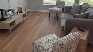 Ett modernt vardagsrum med en grå soffa, en mönstrad pall och en golvlampa. Det finns två fönster med persienner, en tv som är monterad på väggen och ett litet bord med en fjärrkontroll.