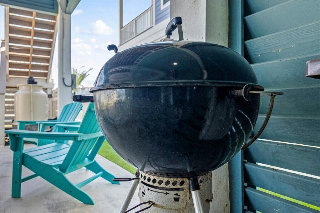 Ein schwarzer Grill mit Deckel, auf einem Gestell platziert, neben einem türkisfarbenen Stuhl und einem weißen Wasserspender.