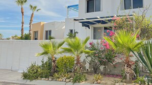 A modern two-story house with a white exterior, a covered patio, and a variety of tropical plants. - Modern 2-Bedroom Condo with A/C in Stunning Los Cabos (MUNICIPIO LOS CABOS)