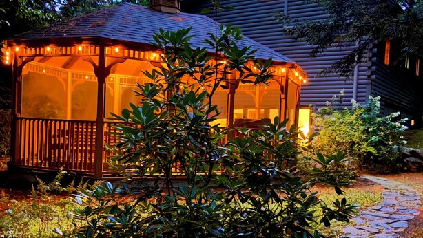 A wooden cabin with a covered porch and string lights, surrounded by dense foliage and a stone pathway.