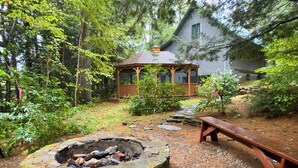 A rustic cabin surrounded by dense forest, a stone fire pit, and a wooden bench.