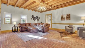 A spacious living room with a brown leather sectional sofa, a small table with a lamp, and a large rug on a brick floor. - Cottage at High Cross Farms | Front Porch (Mount Joy)