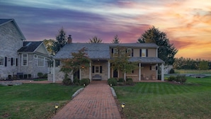 A two-story house with a brick pathway leading to the front door, surrounded by a well-maintained lawn and a stone building to the left. - Cottage at High Cross Farms | Front Porch (Mount Joy)