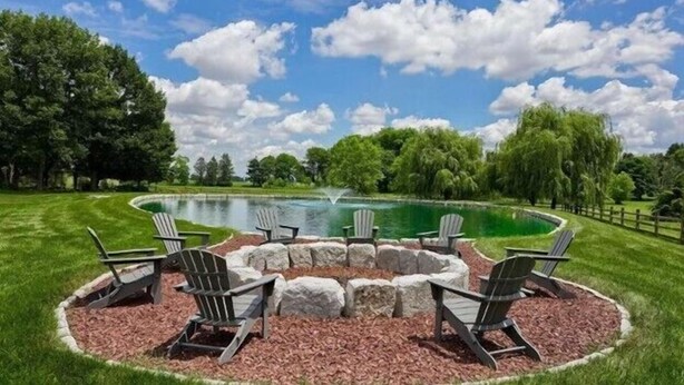 A circular fire pit with four Adirondack chairs around it, set in a grassy area with a pond and trees in the background. - Cottage at High Cross Farms | Front Porch (Mount Joy)