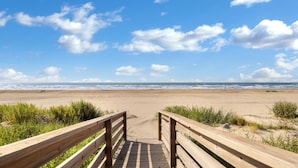 Ein hölzerner Steg führt zu einem Sandstrand, im Hintergrund sind Wellen zu sehen.