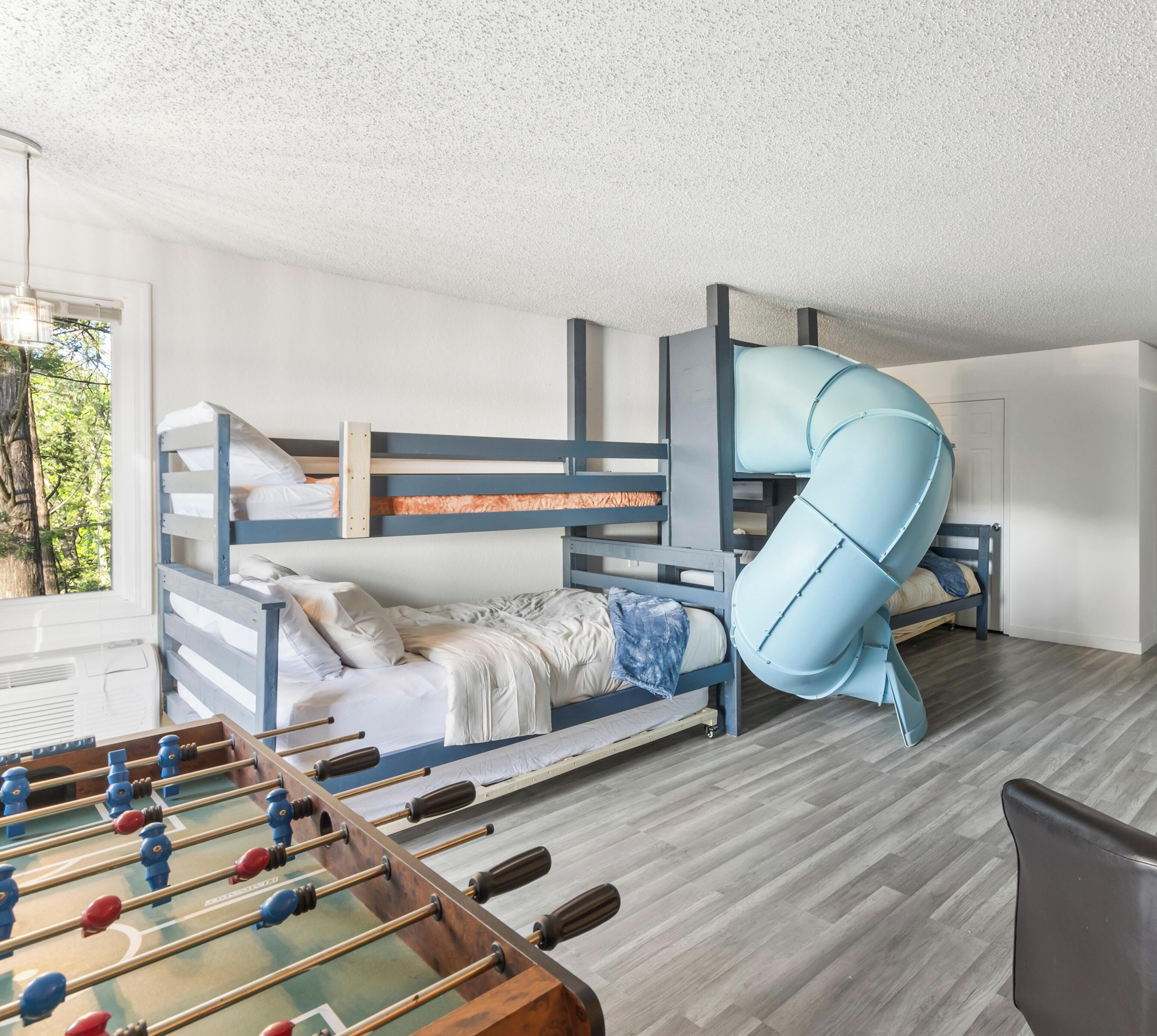 A bunk bed room with a slide, a foosball table, and a window with a view of trees.