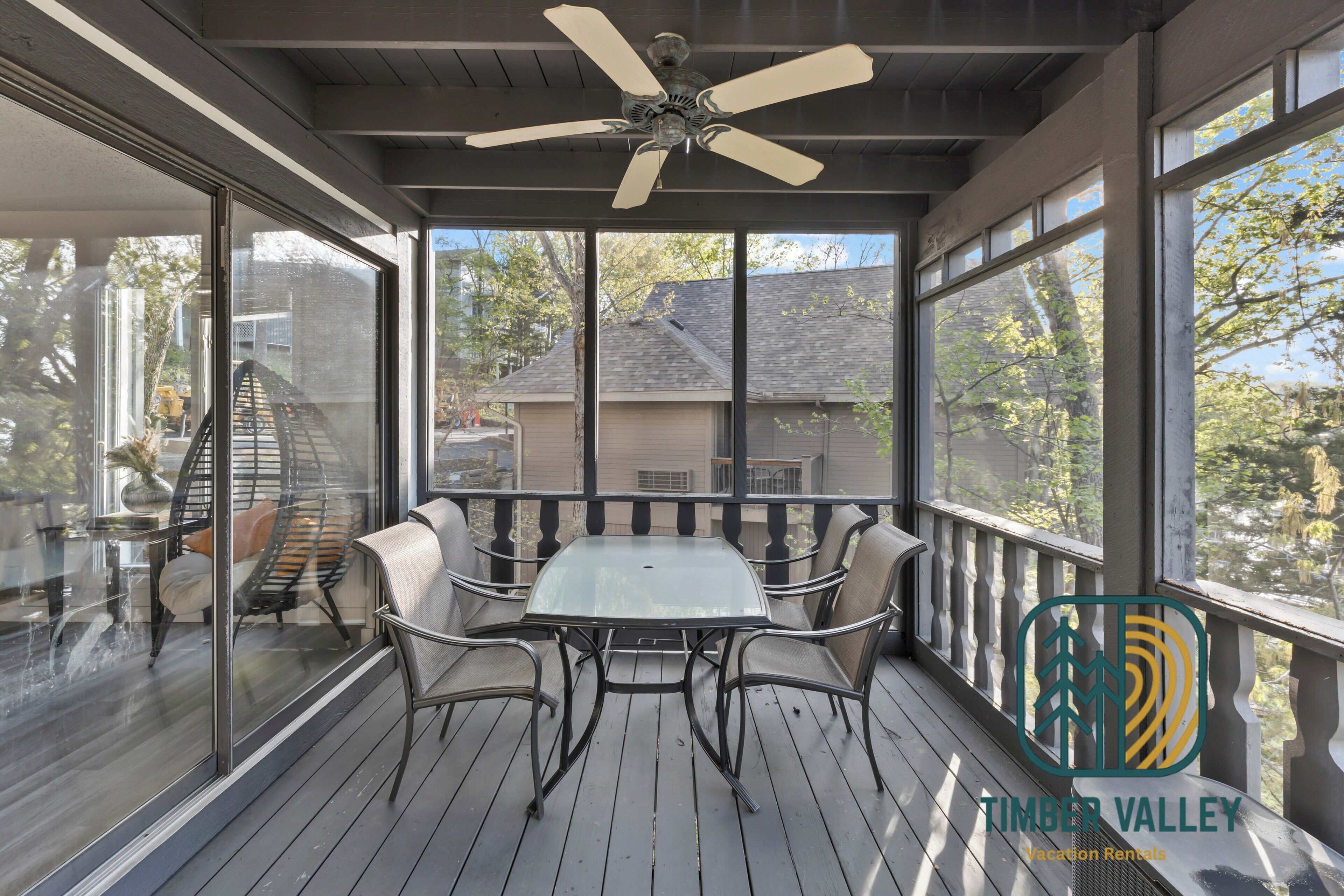 A covered outdoor area with a table and chairs, a ceiling fan, and large windows offering views of a residential area.