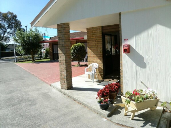 A building with a brick facade, a covered entrance, and a small porch area with potted plants and a white chair.