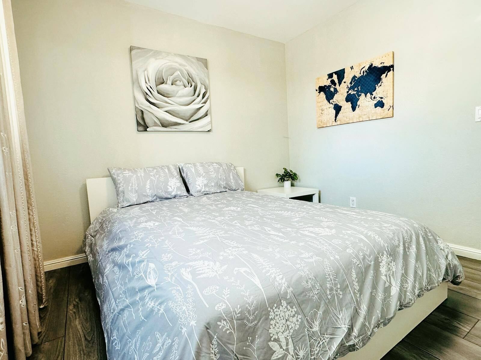 A bedroom with a bed, a nightstand, a potted plant, a world map, and a floral painting.