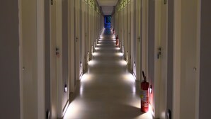 Un largo pasillo de hotel estrecho, con puertas a ambos lados, un extintor en el lado derecho y una zona iluminada con luz azul al final.