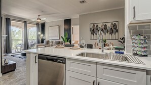 A modern kitchen with a central island, white cabinetry, and a dishwasher. There is a dining area with a sofa and chairs, and a large window with curtains. - Scottsdale 2BR • Pool • Gym •Free Parking •Central (Scottsdale)