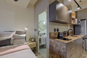 Una camera da letto moderna con un letto grande, un comodino e un tavolino. Accanto alla camera, una piccola cucina abitabile con lavello, fornelli e forno a microonde.