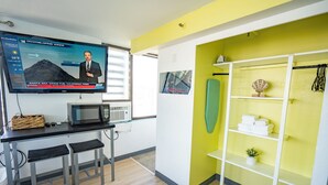 A modern hotel room with a TV displaying a weather forecast, a kitchenette, a yellow accent wall, and a shelf with towels and a decorative item.