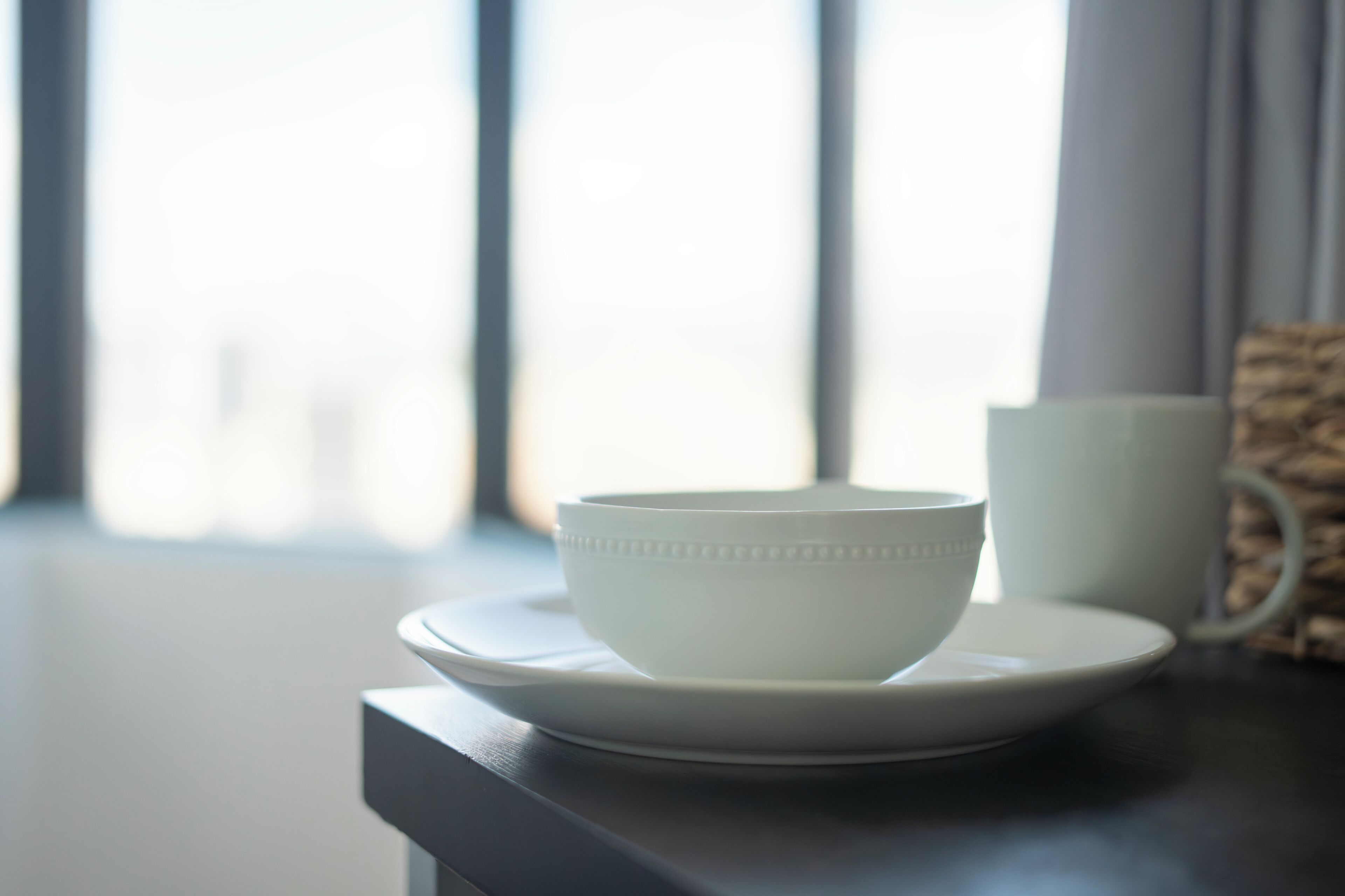 Un bol et une tasse blancs sur une surface sombre, avec une fenêtre en arrière-plan.