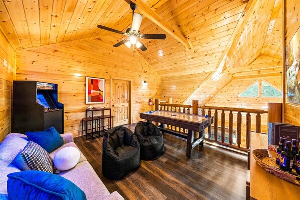 A cozy cabin interior with a wooden ceiling, a ceiling fan, a pool table, a sofa with cushions, and a small bar area with bottles.