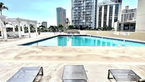 A swimming pool with lounge chairs, surrounded by tall buildings and palm trees.