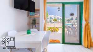 Una habitación moderna con una mesa blanca, sillas, un televisor, una ventana con cortinas amarillas y vistas a una piscina y edificios.