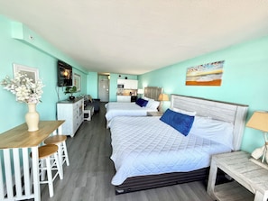 A hotel room with two beds, a kitchenette, a dining area with a table and chairs, and a bathroom visible through an open door.