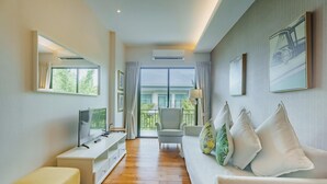 Ein modernes Hotelzimmer mit Holzboden, einem weißen Sofa mit Zierkissen, einem weißen TV-Schrank und einem großen Fenster mit Blick ins Freie.