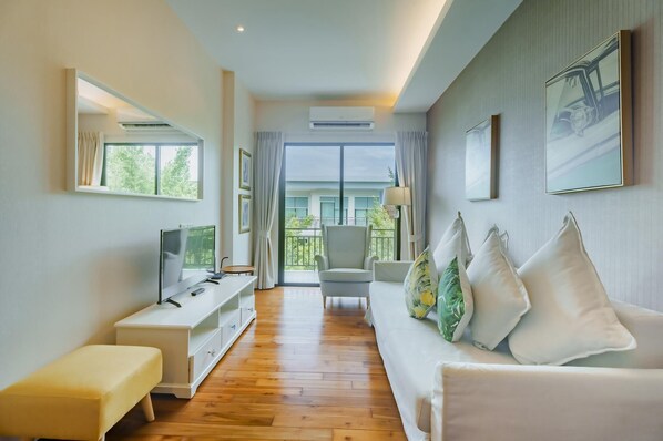 Una habitación de hotel moderna con suelo de madera, un sofá blanco con cojines decorativos, un mueble de televisión blanco y un amplio ventanal con vistas al exterior.
