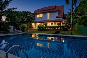A swimming pool with a tiled surface, a house with a red roof, and a well-maintained lawn with various plants. - Koson, Elegant 3 Bedroom Villa (Tambon Rawai)