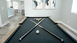 Ein Billardtisch mit einer weißen Spielkugel und einer Reihe Billardkugeln, ein modernes Kunstwerk an der Wand und ein Blick in einen Essbereich.