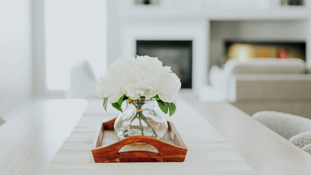 Ein weißer Holztisch in einem modernen Wohnzimmer mit einer weißen Holzschale, einer Glasvase und weißen Blumen.