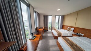 Ein Hotelzimmer mit zwei Betten, einem Schreibtisch mit Stuhl, einem kleinen Tisch ebenfalls mit Stuhl und großen Fenstern mit Vorhängen.
