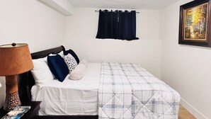 Un dormitorio con cama, mesita de noche con lámpara, alfombra azul estampada, un cuadro en la pared y una luz de techo.