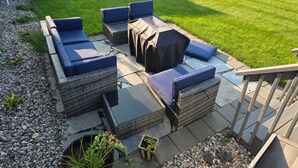 Een terras buiten met rieten meubilair en een overdekte tafel, omringd door een verzorgd gazon en een witte schutting.