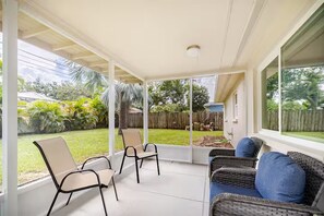 A covered patio with chairs and a wicker sofa, overlooking a backyard with a wooden fence and greenery.