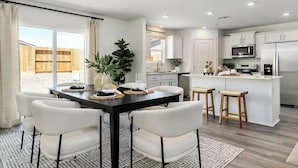 A modern kitchen with white cabinets, a central island with stools, and a dining area with a black table and white chairs. - Large new construction 3-bedroom near Hospital Hub (Bakersfield)