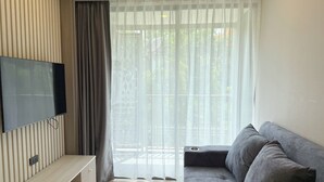 A modern living room with a flat-screen TV mounted on a wall, a grey sofa, and a large window with sheer curtains. - 1BR Condo Near Beach with Pool Gym C235 (Choeng Thale)