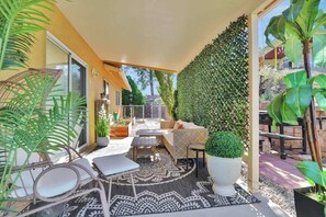Un espace extérieur ensoleillé, aménagé avec des meubles en osier, un grand tapis à motifs et une grande plante en pot.