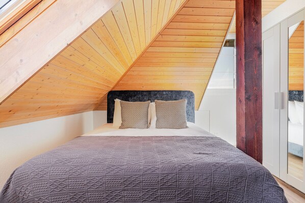 Un dormitorio con techo inclinado, vigas de madera, una cama con ropa de cama estampada y vistas a través de un ventanal.