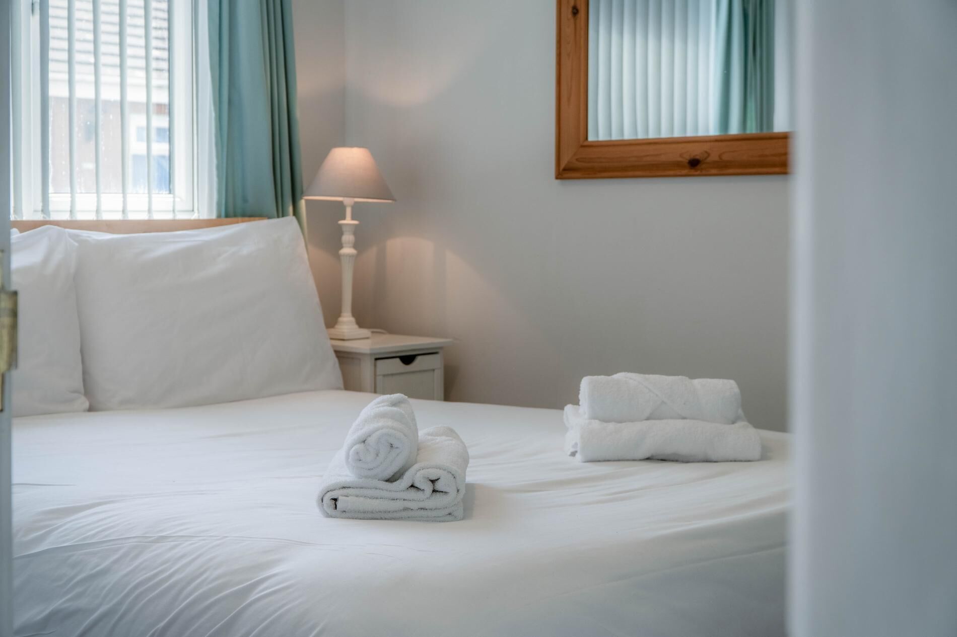 A neatly made bed with white linens, a rolled towel, and a folded towel on a white nightstand beside it.