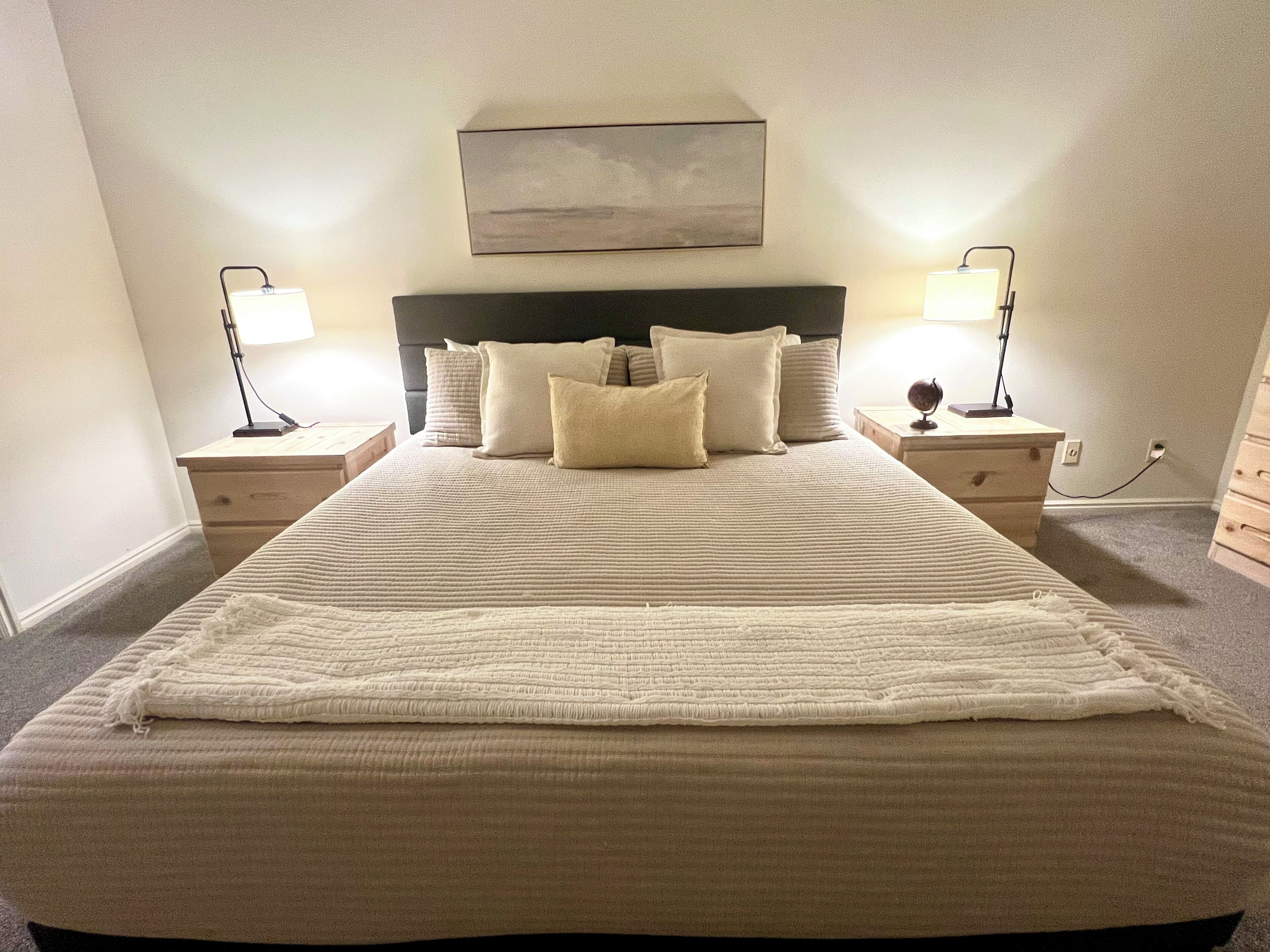 Un lit bien fait, recouvert d’une courtepointe blanche à motifs, et agrémenté de plusieurs oreillers, avec de chaque côté des tables de chevet surmontées de lampes.