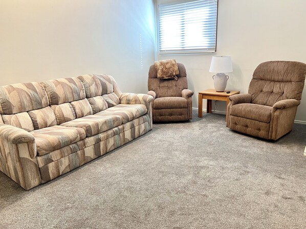 A living room with a striped sofa, a recliner, and a chair, a small table with a lamp, and a window with blinds.