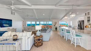 Un vaste espace de vie avec une grande fenêtre donnant sur l’océan, une cuisine dotée de meubles blancs et un coin repas avec des chaises blanches.
