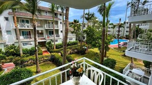 A balcony with a table and flowers, overlooking a pool and a well-maintained garden with palm trees and a multi-story building in the background. - Sol Caribe Primavera Pool (Punta Cana)