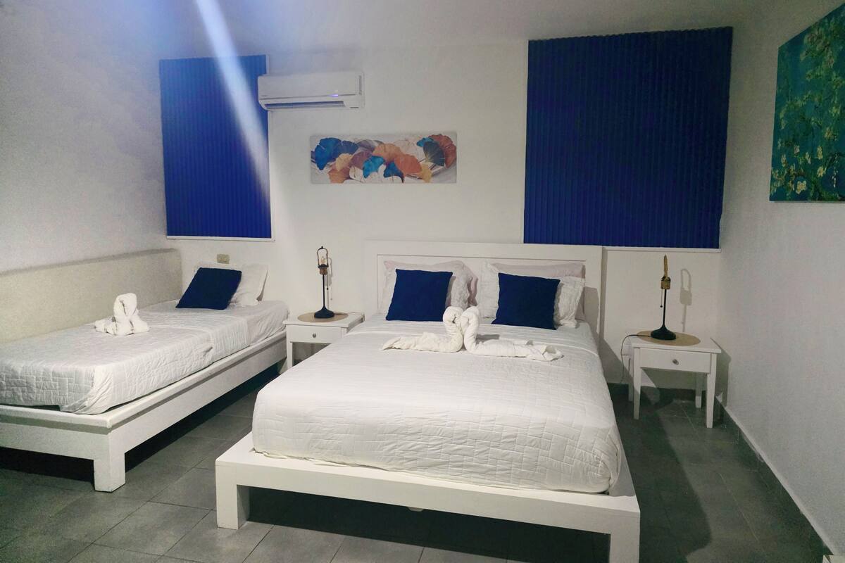 A hotel room with two single beds, a large bed, a wall-mounted air conditioner, a painting, and a blue curtain.