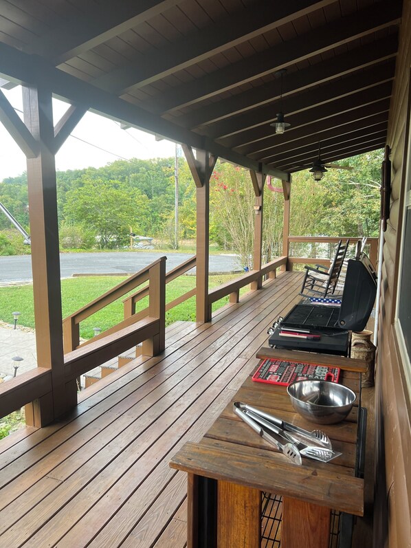 Een overdekt terras buiten met een barbecue, kookgerei en uitzicht op een meer en bos.
