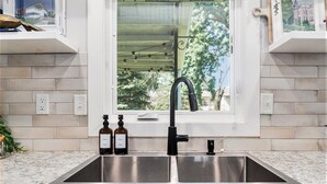 Una cocina con un lavamanos doble, dos botellas de jabón para manos y una ventana que ofrece vistas a árboles y un edificio.