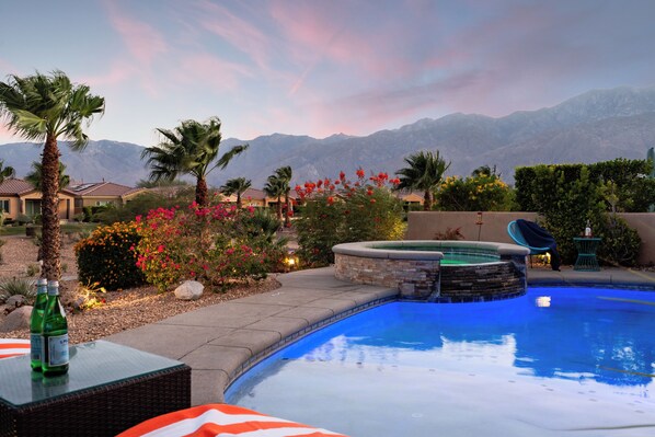 A swimming pool with a hot tub, surrounded by palm trees and flowering plants, with mountains in the background.