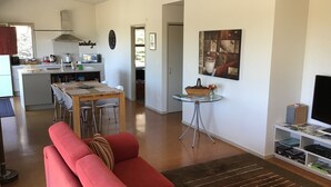 A modern living room with a red sofa, a dining area with a wooden table, a kitchen with white cabinets, and a TV on a stand. - Ready for a cosy Beach holiday? (Foxton Beach)